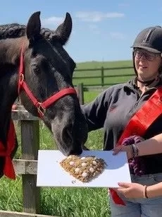 Black horse Maya is wearing a red birthday sash and is tucking into a horse-friendly cake, supported by two of her carers also wearing red sashes.