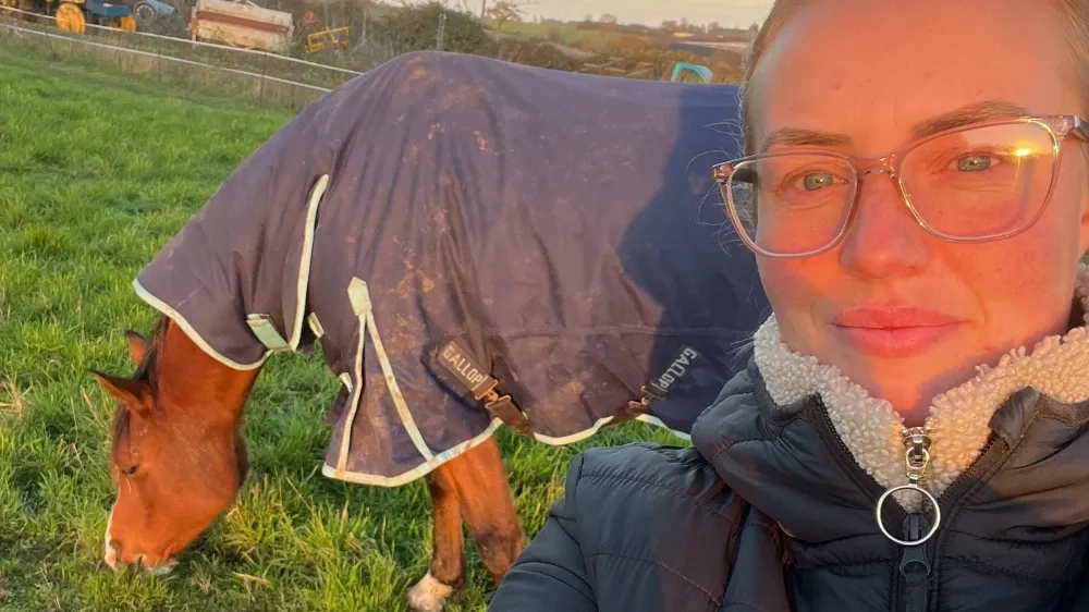 Photo of a horse wearing a rug out eating grass (Zuri, left) and their owner Amy (right).