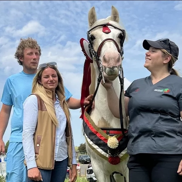 A brown and white pony wearing a winner's sash stands next to Redwings vet Nicola and their owners.